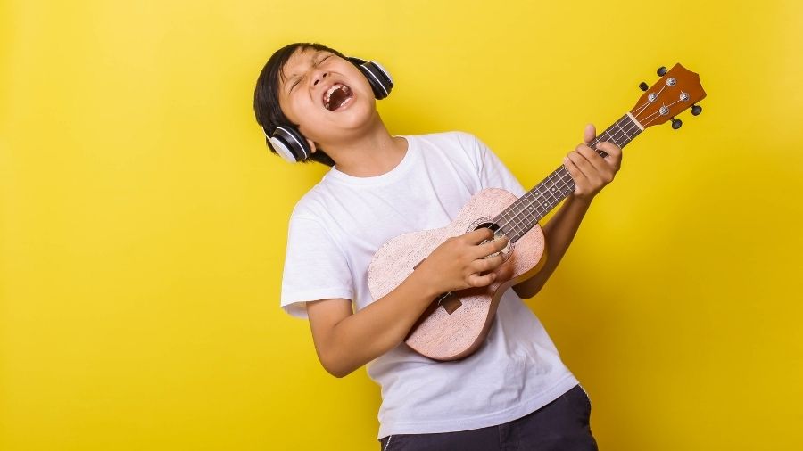 oy singing and playing a ukulele while wearing a headset.