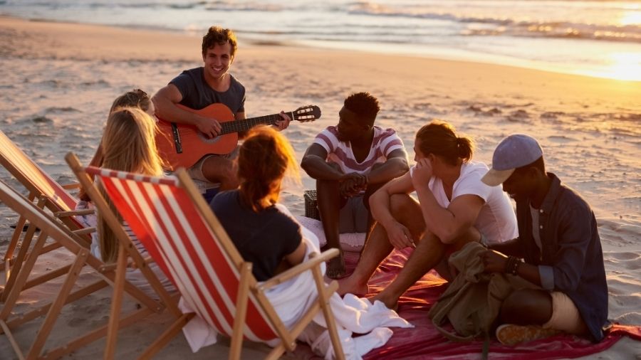 A group of friends sitting in a circle on the beach, with one person playing a guitar.