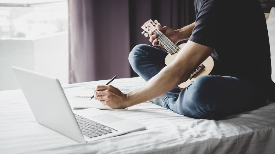 Songwriter Composing Music with Laptop and Ukulele on Bed