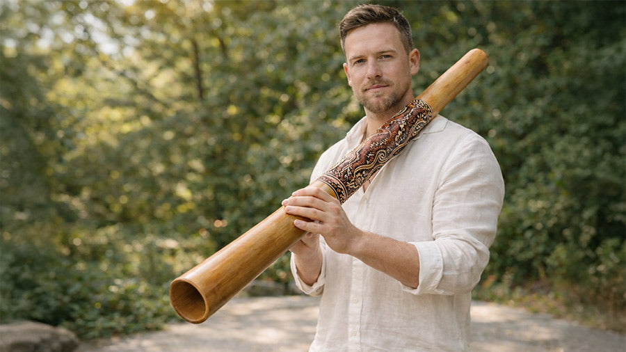 A well-groomed man in a white linen shirt holding a traditional bamboo didgeridoo outdoors, showing confident playing posture, clean natural wood finish, and detailed hand-painted artwork against a soft green background.