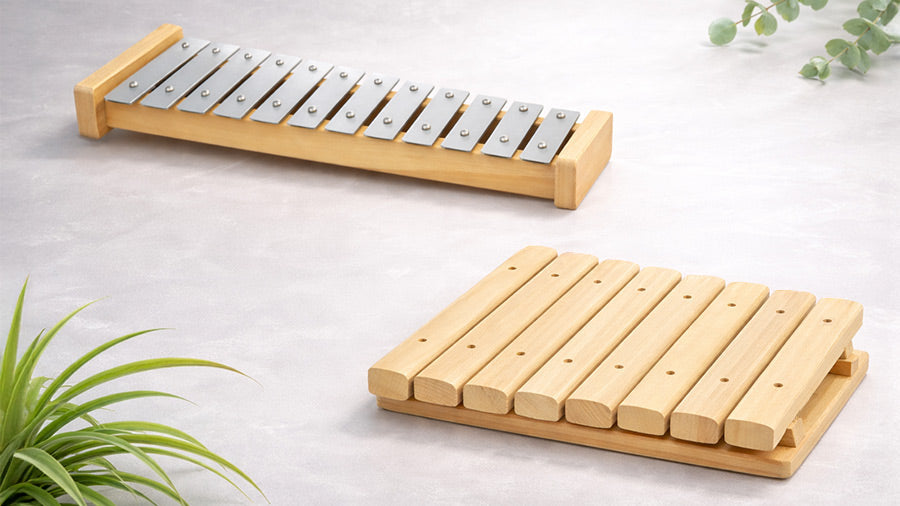 Minimal studio image of a metal bar xylophone and a wooden xylophone on a soft textured light background, with subtle eucalyptus and spider plant foliage in the corners, styled in a clean Apple-inspired aesthetic.