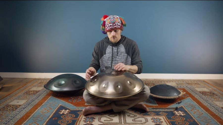 A man sitting on a carpet playing a nitrided steel, tongue drum and a stainless steel drum, with the instruments placed in front of him.