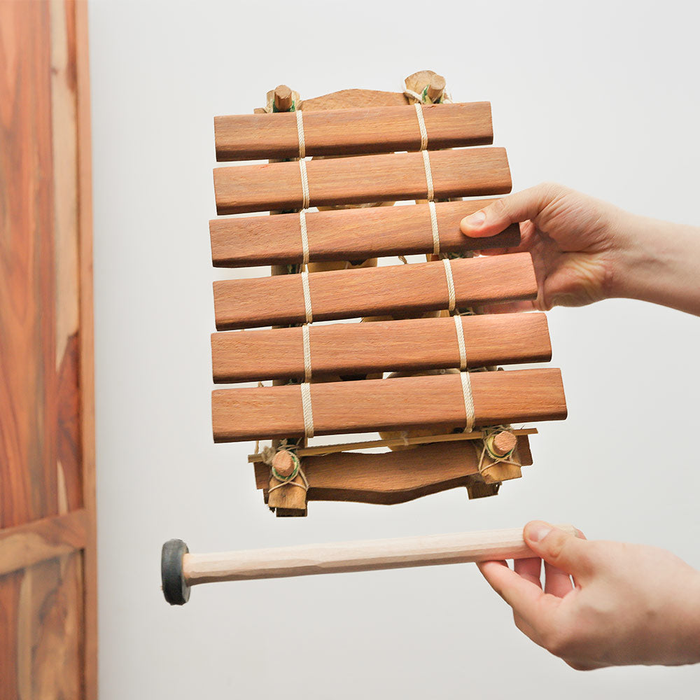 A person holding a handmade balafon xylophone with wooden keys and gourd resonators, alongside two mallets.