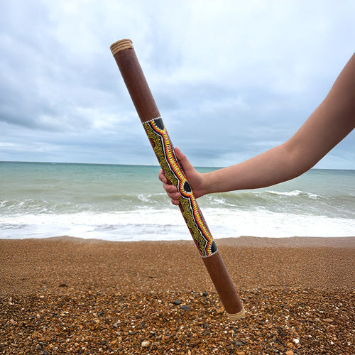 Hand holding a decorated rainstick on a beach with ocean in the background