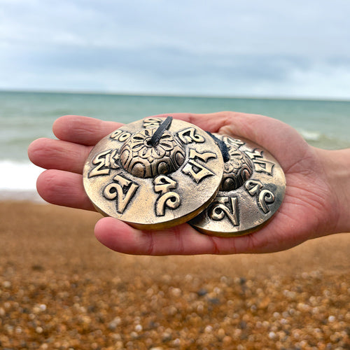 Hand holding two brass bells with intricate designs on a beach.