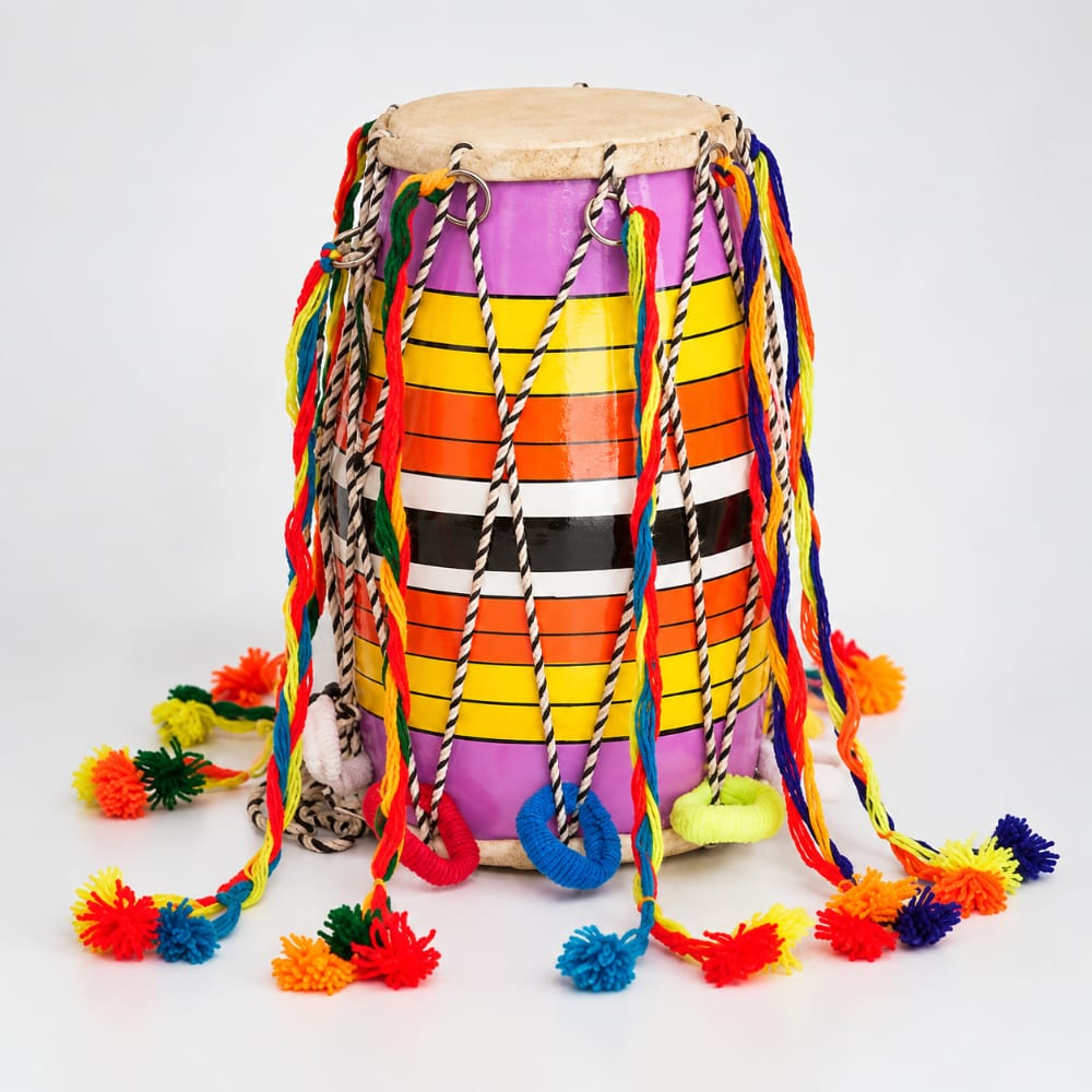 A colorful bhangra dhol drum with a purple and orange striped body, a natural drum head, and numerous multicolored hanging cords and pom-poms, photographed on a clean grey studio background.