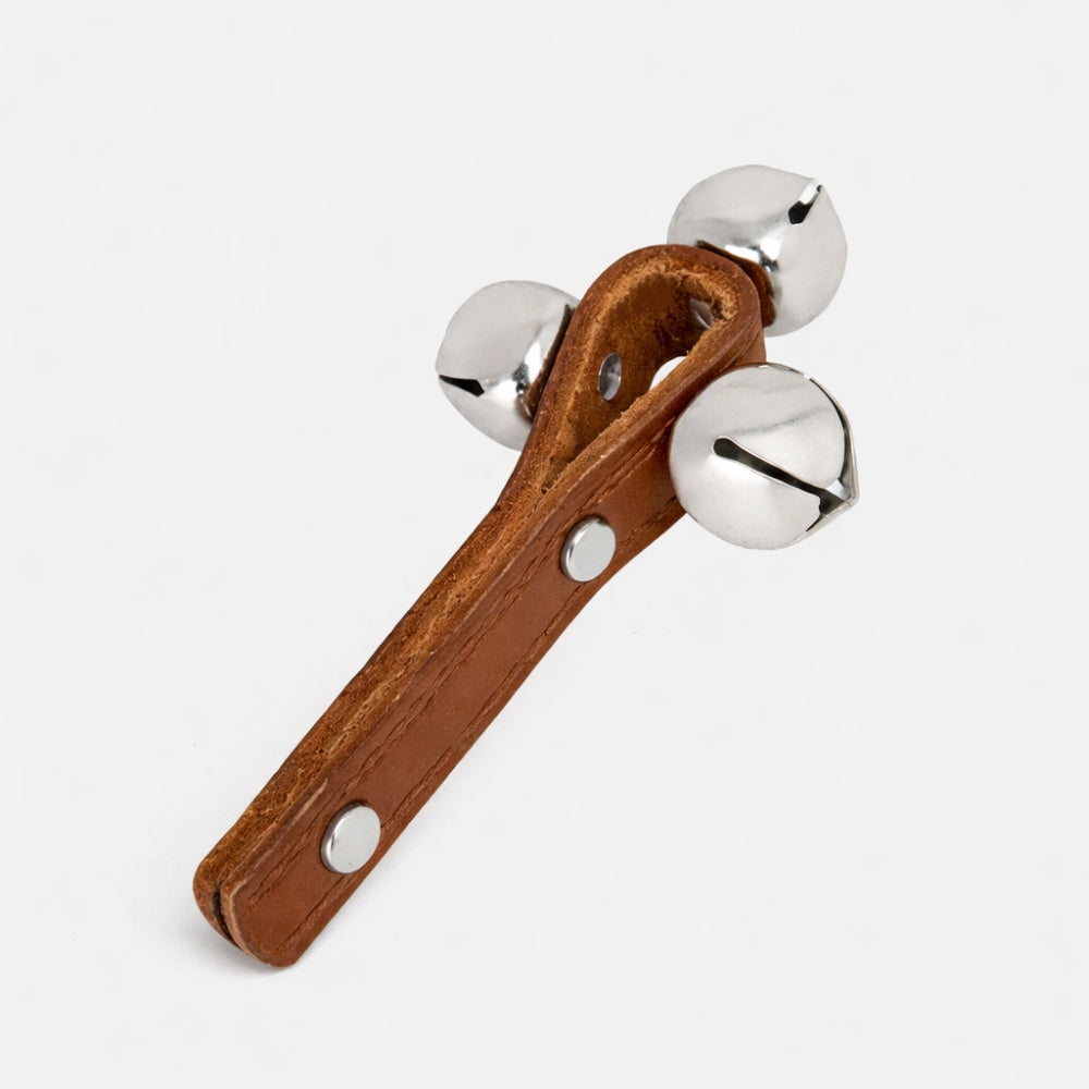Wooden jingle bell instrument with three silver bells attached to a leather strap on a white studio background
