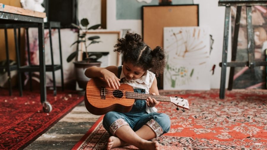 A young girl in a denim jumper sitting on the floor while playing a ukulele.