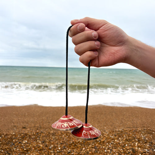 Hand holding two red and silver bells on strings against a beach background