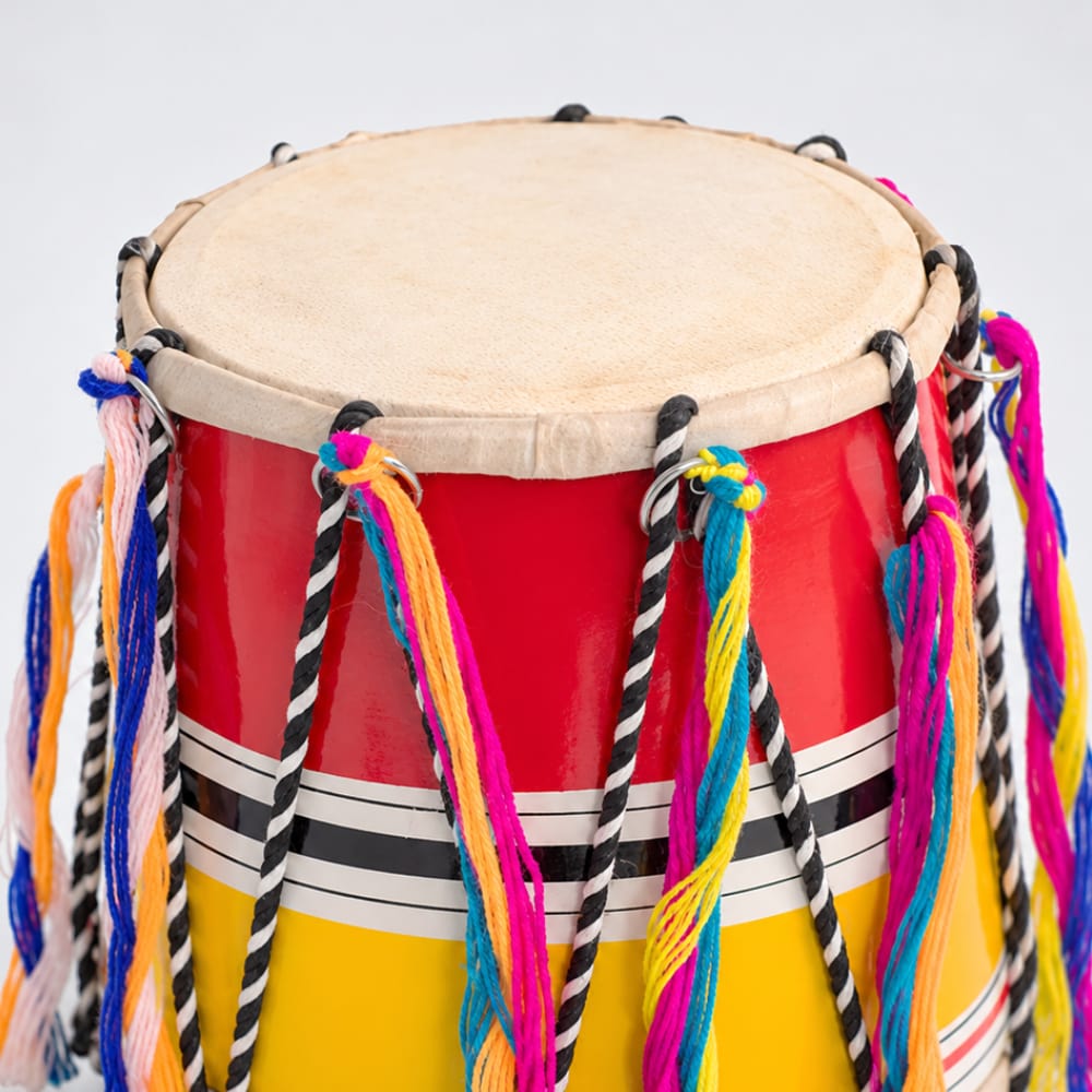 Close up of the colourful bhangra dhol drum with a natural beige drumhead, a red upper body, a yellow lower body, and multicolored hanging tassels against a light gray background.