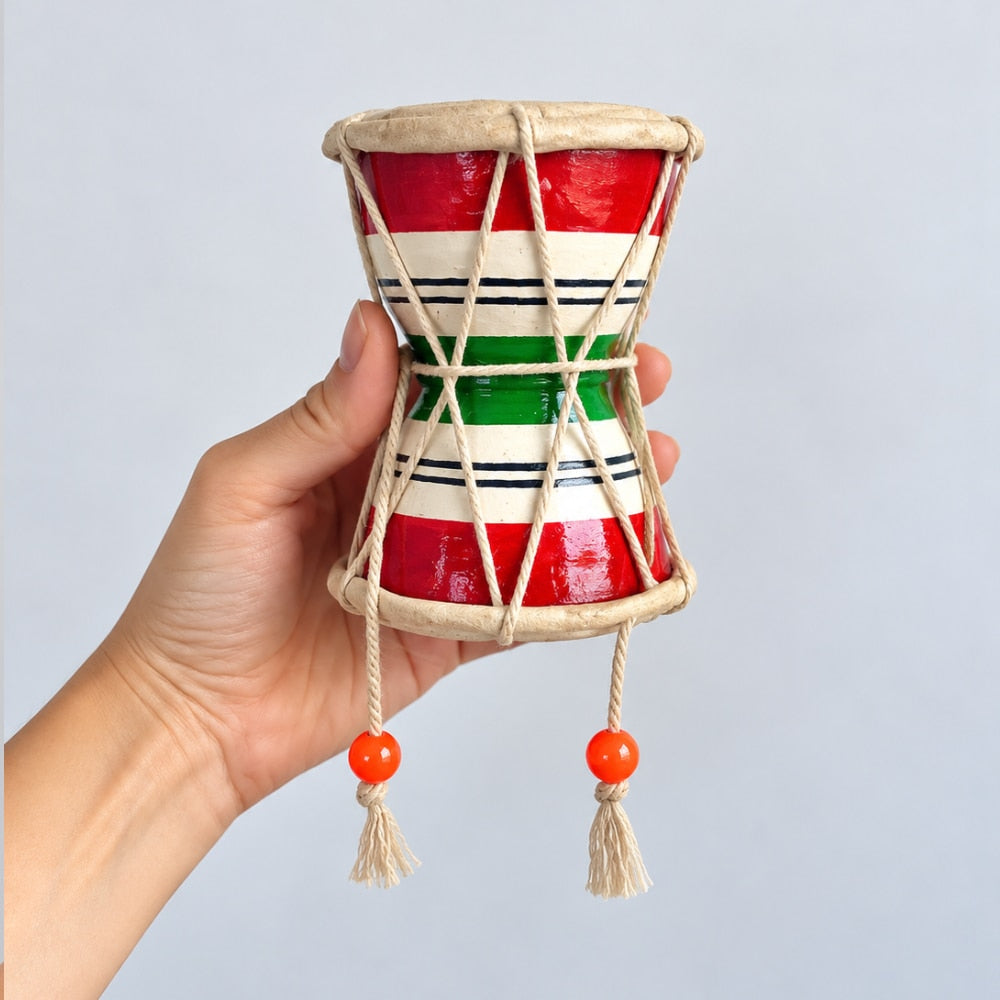 A hand holding a small hand drum with a red, white, and green striped body, beige rope tensioning, and orange bead tassels, set against a plain light gray background.