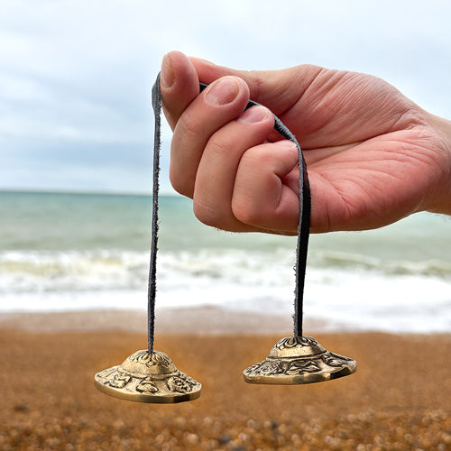 Hand holding two ornate brass tingsha on a beach