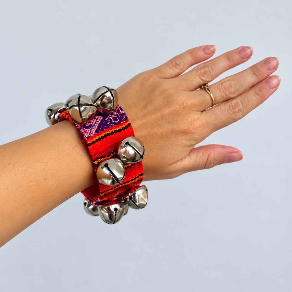 A close-up of a person's hand and wrist wearing a stack of colorful woven bracelets adorned with silver bells, set against a plain light gray background.