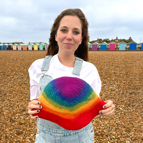 Woman holding a rainbow-colored knitted hat on a beach with colorful huts in the background