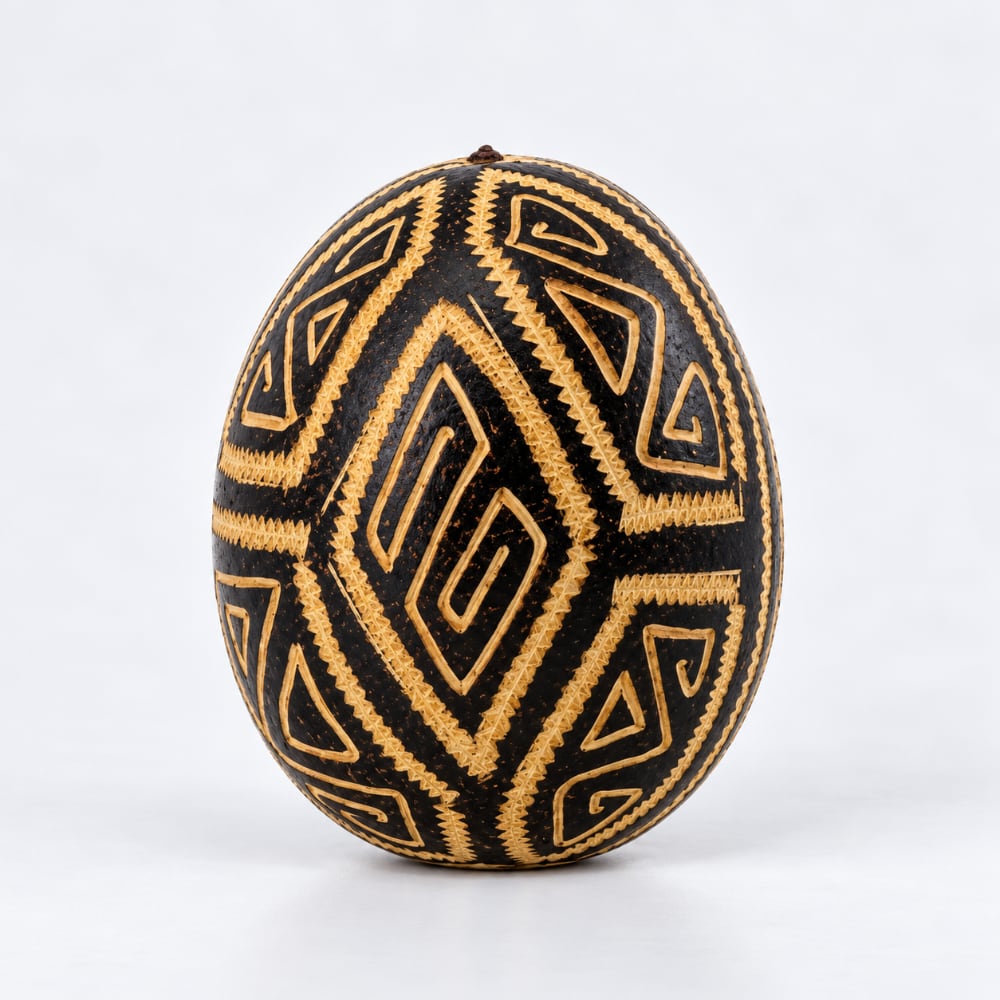A handcrafted tribal Shipibo egg with intricate geometric patterns in black and natural tan wood, photographed on a light grey studio background.