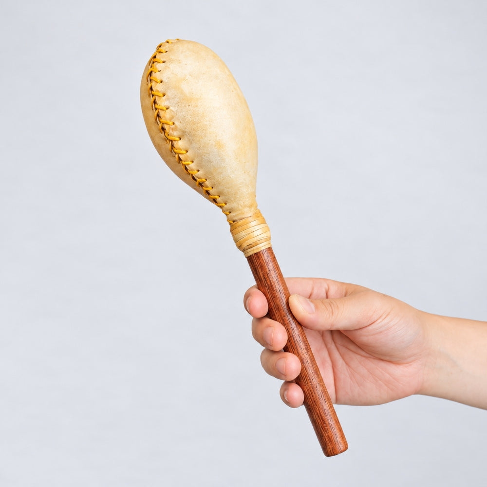 A hand holding a terre shamanic shaker with a light beige stitched animal hide head and a brown wooden handle set against a plain light gray studio background.